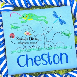 May include: A light blue t-shirt featuring a green frog leaping over a black line with a red ladybug, green crickets, and a blue dragonfly. The shirt has green grass at the bottom and the text "Sawyers Charm Embroidery Designs" and "Cheston" in blue.