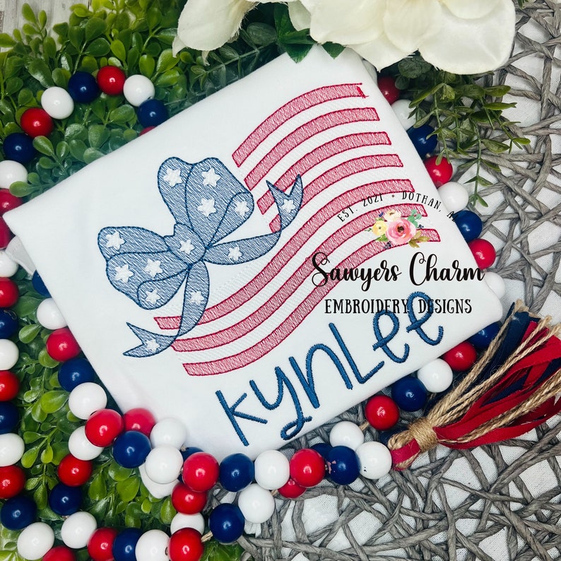 May include: A white t-shirt with a red, white, and blue embroidered American flag design with a bow. The name "Kynlee" is embroidered below the flag. The text "Sawyers Charm Embroidery Designs" is embroidered below the name.