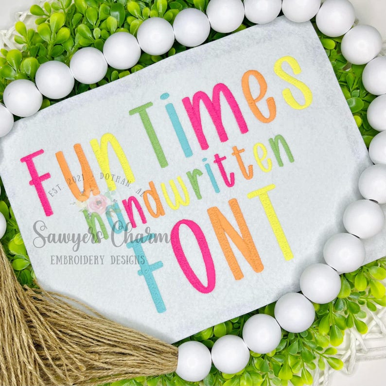 May include: A white tag with the words "Fun Times Handwritten Font" in colorful, playful lettering. The tag is surrounded by greenery, white beads, and a jute tassel. The text includes the words "Sawyers Charm Embroidery Designs".