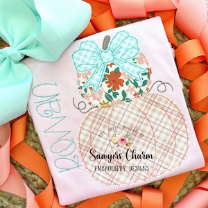May include: A pink shirt with a stitched design of two pumpkins. The top pumpkin has a floral pattern and a blue and white gingham bow. The bottom pumpkin has a plaid pattern and the text "Sawyers Charm Embroidery Designs" and "Est. 2021 Dothan, AL".