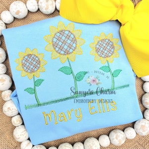 Puede incluir: Una camiseta azul claro con tres girasoles bordados y el texto "Mary Ellis" en hilo amarillo. El texto "Sawyers Charm Embroidery Designs" también está bordado en hilo verde.