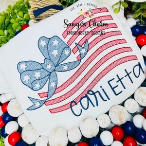 May include: White t-shirt with red, white, and blue embroidered American flag design with a bow and the name "Carletta" embroidered in blue.