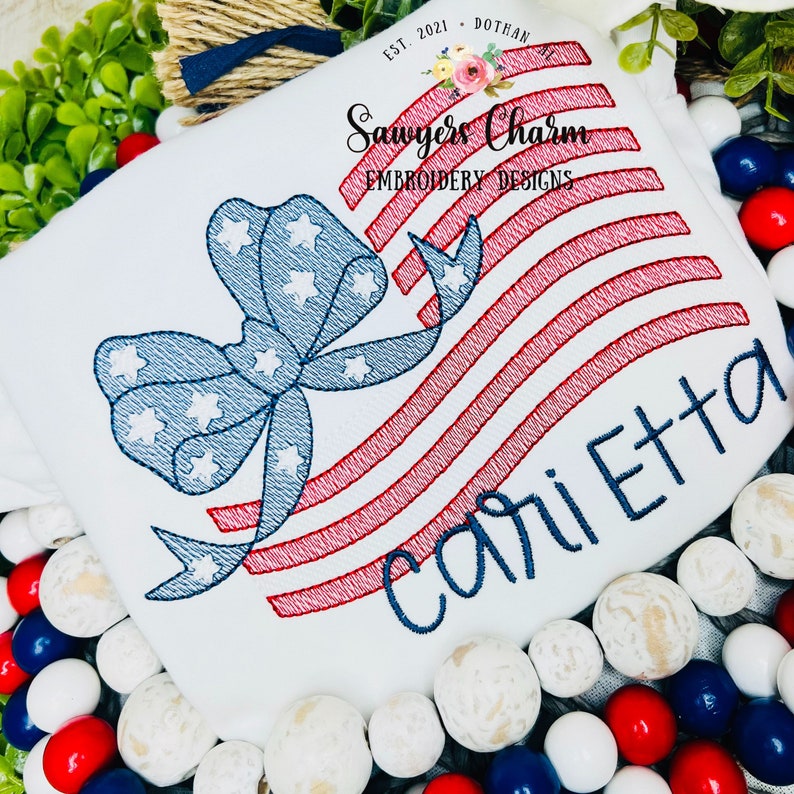 May include: White t-shirt with red, white, and blue embroidered American flag design with a bow and the name "Carletta" embroidered in blue.