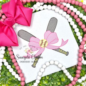 May include: A pink bow and a baseball-themed embroidery design. The design features crossed baseball bats, a baseball, and a pink bow. The text "Sawyers Charm Embroidery Designs" is also visible.