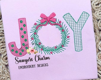 Op de afbeelding: Roze en groene geborduurde afbeelding van het woord "JOY" met een krans en een strik. Het ontwerp is op een roze shirt met de tekst "Sawyers Charm Embroidery Designs".