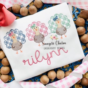 May include: A white onesie with three embroidered turkeys in blue, pink, and green plaid. The turkeys have brown bodies and are wearing bows. The onesie is personalized with the name "Rilynn" in pink cursive lettering. The text "Sawyers Charm Embroidery Designs" is also embroidered on the onesie.