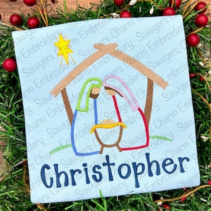 May include: Embroidered design of the nativity scene with a manger, Mary, Joseph, and baby Jesus. The design is on a light blue fabric with the name "Christopher" embroidered below.