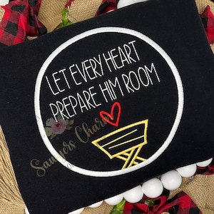 May include: Black sweatshirt with the embroidered phrase "LET EVERY HEART PREPARE HIM ROOM" in a white circle. A red heart and a gold manger are below the text. The sweatshirt is surrounded by a decorative wreath.