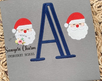 May include: A gray t-shirt with a blue embroidered letter A and two red and white embroidered Santa faces. The text "Sawyers Charm Embroidery Designs" is embroidered below the letter A. The text "Est. 2021 Dothan, AL" is embroidered above the letter A.