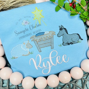 May include: A blue baby onesie with a white embroidered design of a manger scene with a donkey, a sheep, and a star. The onesie also has the name "Rylee" embroidered on it.