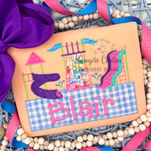 May include: A peach-colored t-shirt with an embroidered design of a playground with a slide, a climbing structure, and a sandbox. The name "Blair" is embroidered in pink letters below the playground design.