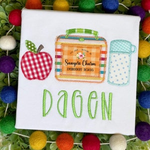 Könnte beinhalten: Ein weißes T-Shirt mit einem grünen gestickten Namen "Dagen" und einem rot-weiß karierten Apfel, einer Lunchbox mit einem Etikett, auf dem "Sawyer's Charm Embroidery Designs" steht, und einer blau-weiß gepunkteten Wasserflasche.