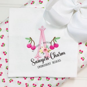 May include: A pink embroidered letter A with cherry blossoms and two cherries on either side. The text "Sawyers Charm Embroidery Designs" is below the letter. The text "EST 2021 DOTHAN AL" is to the left of the letter.
