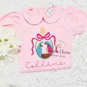 May include: Pink short-sleeved shirt with a Peter Pan collar. The shirt features an embroidered nativity scene with a star, bow, and the name "Collins" in cursive. The design includes blue, pink, and gold colors.