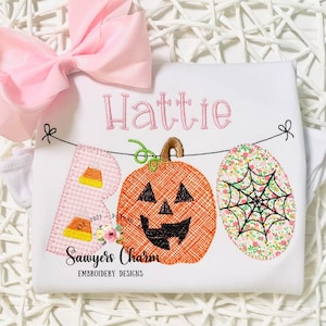 May include: A white onesie with the name "Hattie" embroidered in pink. The onesie features a pink, orange, and green embroidered BOO banner with a pumpkin, candy corn, and spiderweb design. The text "Sawyers Charm Embroidery Designs" is also embroidered on the onesie.