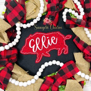 Può includere: Una ghirlanda decorativa con un centro in feltro nero, un disegno di maiale rosso e il nome "Ellie" in scrittura bianca. È adornata con fiocchi a quadri di bufalo rossi e neri, juta e perline di legno bianche. Il testo "Sawyers Charm" è anche visibile.