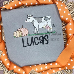 以下が含まれることがあります： 牛、カボチャ2つ、そして「Lucas」という名前が刺繍されたグレーのTシャツです。