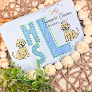 May include: A light blue shirt with the embroidered letters H, L, and S in a light blue color with a green outline. Two embroidered yellow puppies are on the shirt. The text "Sawyers Charm Embroidery Designs" is on the shirt.