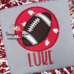 Op de afbeelding: Een grijs T-shirt met een rode cirkel met daarin een bruine voetbal en witte olifanten. De tekst "Sawyers Charm Embroidery Designs" en "EST. 2021" staan in de linkerbovenhoek. De naam "LUKE" is in rood onder de cirkel geborduurd.