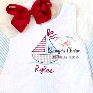May include: White seersucker dress with a sailboat design. The sailboat has red and blue striped sails and a gray checkered hull. The name "Rylee" is embroidered in burgundy. A large red bow is at the top.