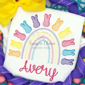 Puede incluir: Camiseta blanca con un diseño de arcoíris y conejitos de colores. La camiseta presenta un arcoíris con conejitos bordados en varios colores. El nombre "Avery" está escrito en rosa debajo del arcoíris. Un lazo amarillo y una cinta morada están en el fondo.