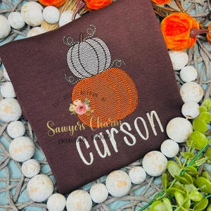 以下が含まれることがあります： オレンジとグレーの2つのカボチャの刺繍デザインが施された茶色のTシャツ。花のアクセント付き。「Carson」という名前が白い筆記体で刺繍されています。「Sawyers Charm」と「Est. 2021」の文字も表示されています。