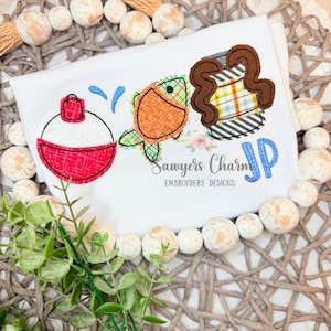 May include: White t-shirt with embroidered fishing-themed designs. Features a red and white bobber, an orange fish with green plaid fins, and a brown and plaid tackle box. The text "Sawyers Charm Embroidery Designs" and the initials "JP" are also embroidered.