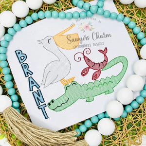 May include: Embroidered design featuring a pelican, crawfish, and alligator. The name "BRANT" is embroidered in blue. The design is on a white background, surrounded by a beaded garland and a woven base.