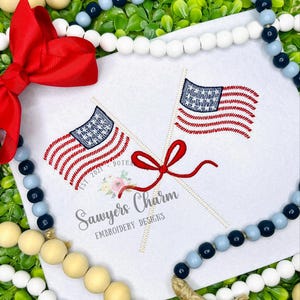 May include: A white square with two embroidered American flags, red and blue bow, and text "Sawyers Charm Embroidery Designs". The flags have red and white stripes and a blue field of stars. Decorative wooden beads and greenery surround the square.