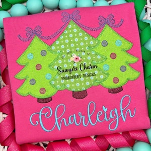 May include: A pink shirt with a design of three green Christmas trees with colorful ornaments and bows. The trees are decorated with white polka dots. The text "Sawyers Charm Embroidery Designs" is on the shirt. The name "Charleigh" is embroidered in blue on the bottom of the shirt.