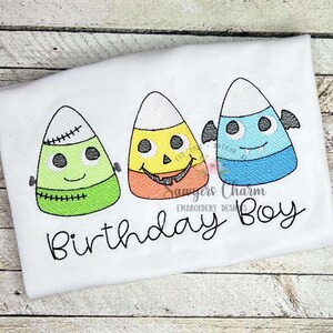 May include: White t-shirt with embroidered candy corn characters: a Frankenstein, a jack-o'-lantern, and a vampire. Each has a smiling face. The text "Birthday Boy" is written in cursive below the candy corn designs. A fun Halloween theme.