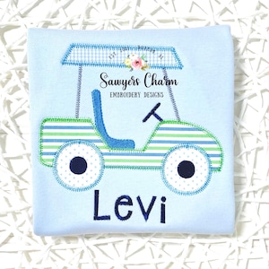 May include: Pale blue t-shirt featuring an embroidered golf cart design. The golf cart has blue and green stripes, a blue seat, and a gingham roof. The name "Levi" is embroidered in navy blue below the cart. The text "Sawyers Charm Embroidery Designs" is also visible.