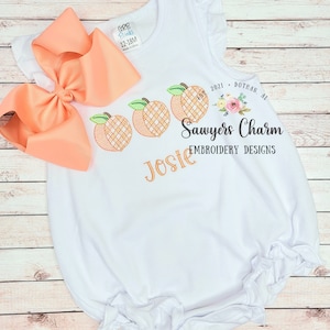 May include: White baby onesie with a peach design and the name "Josie" embroidered on the front. The onesie also has the text "Sawyers Charm Embroidery Designs" and "2021 Dothan, AL" embroidered on the front.