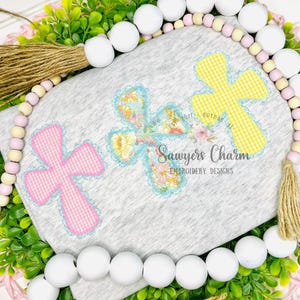 May include: A gray fabric with three embroidered crosses in pink gingham, floral, and yellow gingham patterns. The design includes the text "Sawyers Charm Embroidery Designs" and is surrounded by a decorative beaded garland and greenery.