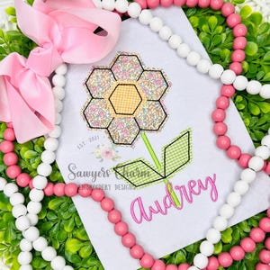 May include: A white card with a floral design and the name "Audrey" in pink script. The flower has a yellow center and pink and yellow petals. A pink bow and pink and white beads surround the card. The text "Sawyers Charm Embroidery Designs" is also visible.