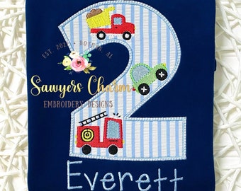 Peut inclure: Un t-shirt rayé bleu et blanc avec un grand numéro deux brodé dessus. Le numéro deux est décoré de trois véhicules jouets : un camion de pompiers rouge, une voiture verte et un camion à benne basculante jaune. Le t-shirt a également le texte "Sawyers Charm Embroidery Designs" et "Everett" brodé dessus.