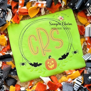 May include: A green t-shirt with a black spiderweb and bats embroidered on it. The center of the web has a monogram "CRS" in orange and black. A smiling orange pumpkin is below the monogram. The text "Sawyer's Charm Embroidery Designs" is above the web.