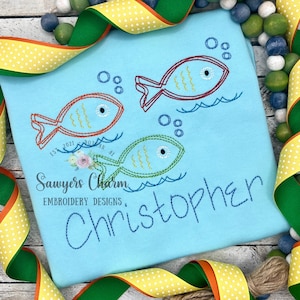 May include: A light blue embroidered t-shirt with three colorful fish and the name "Christopher" stitched in blue thread. The fish are surrounded by bubbles and waves.