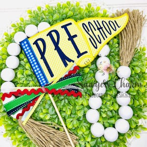 May include: A decorative preschool-themed flag with the words "PRE SCHOOL" in navy blue on a yellow gingham background. The flag is attached to a wooden stick and adorned with colourful ribbons and a jute tassel, set against a green wreath.