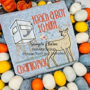 May include: Embroidered design on a gray t-shirt featuring a deer, a hunting blind, and the text "teach a boy to hunt". The text "Sawyers Charm Embroidery Designs" and "Charlie Mac" are also included.