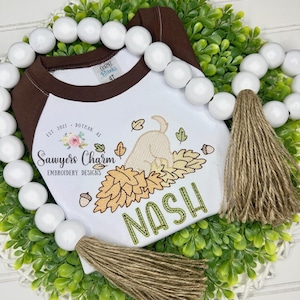 May include: White and brown raglan style shirt with an embroidered design. The design features a dog playing in leaves, acorns, and the name "NASH" in green. The shirt also has the text "Sawyers Charm Embroidery Designs" and "Est. 2021 Dothan, AL".