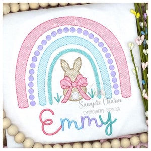 May include: White t-shirt with an embroidered rainbow design in pink, blue, and purple. A bunny with a pink bow is centered under the rainbow. The name "Emmy" is embroidered in pink and blue script below the rainbow.