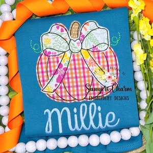 Pode incluir: Uma camisa azul-petróleo com uma aplicação de abóbora com um padrão xadrez e laço floral. O nome "Millie" é bordado abaixo. A camisa é decorada com fita laranja e contas brancas.