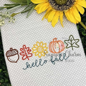 Könnte beinhalten: Gesticktes Herbstmotiv auf hellgrauem Stoff. Mit Eichel, Blatt, Sonnenblume, Kürbis und Blatt. Die Worte "hello fall" sind in Schreibschrift gestickt. Eine Sonnenblume und Grünzeug im Hintergrund.