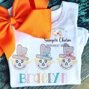 May include: White sleeveless shirt with three embroidered scarecrows and the name "Braelyn" in colorful letters. The shirt has a small logo that says "Sawyers Charm Embroidery Designs" with the text "Est. 2021 Dothan, AL".