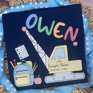 May include: A blue embroidered design featuring the name "Owen" in rainbow colors, a yellow construction crane lifting a stack of papers, a blue backpack, pencils, and an apple. The design includes the text "Sawyer's Charm Embroidery Designs".