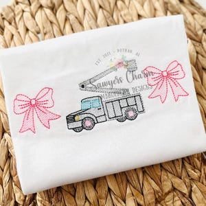 May include: A white t-shirt with an embroidered design of a pink bow, a gray truck with a lift, and the text "Sawyers Charm Embroidery Designs Est 2021 Dothan, AL".