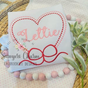 May include: A white onesie with pink and red embroidered hearts and a red bow. The onesie is personalized with the name "Lettie" and the text "EST. 2024 - DOTHAN, AL" and the text "Sawyers Charm EMBROIDERY DESIGNS".