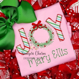 May include: Pink Christmas-themed shirt with the word "JOY" embroidered in candy cane stripes and a green wreath with a red bow. The name "Mary Ellis" is embroidered below the word "JOY".