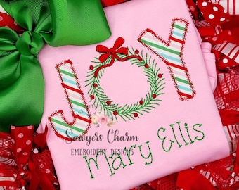 Op de afbeelding: Roze kerst-shirt met het woord "JOY" geborduurd in candy cane strepen en een groene krans met een rode strik. De naam "Mary Ellis" is onder het woord "JOY" geborduurd.